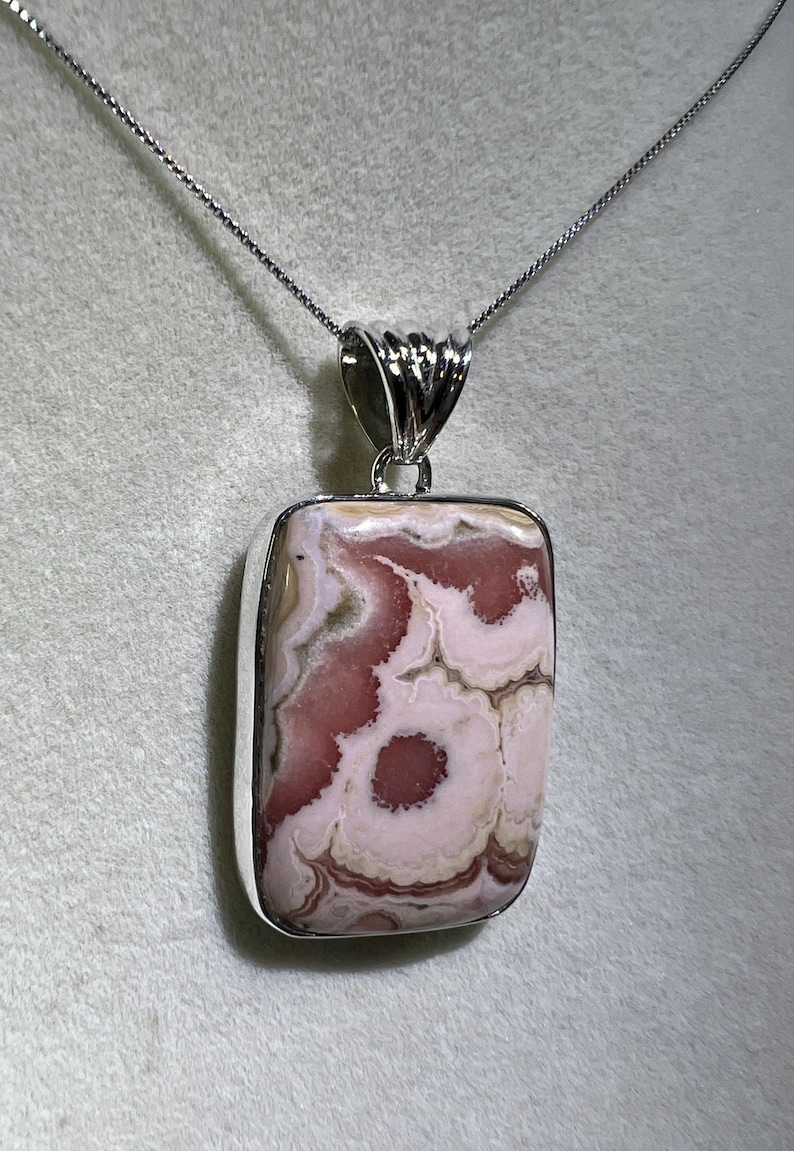 May include: A rectangular pendant with a silver-colored bezel and chain. The pendant features a polished stone with a pattern of pink and white swirls. The stone has a unique, natural appearance, and the chain is delicate.