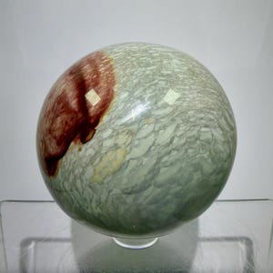 May include: A polished stone sphere with a mottled green and grey surface, featuring a large, reddish-brown patch. The sphere is displayed on a clear glass stand, highlighting its natural patterns and colours. This decorative item is ideal for home decor.