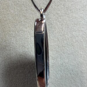 May include: A silver-toned pendant with a dark, reflective center. The pendant has a rounded rectangular shape with a loop at the top for a chain. The edges are polished, creating a contrast with the darker interior.