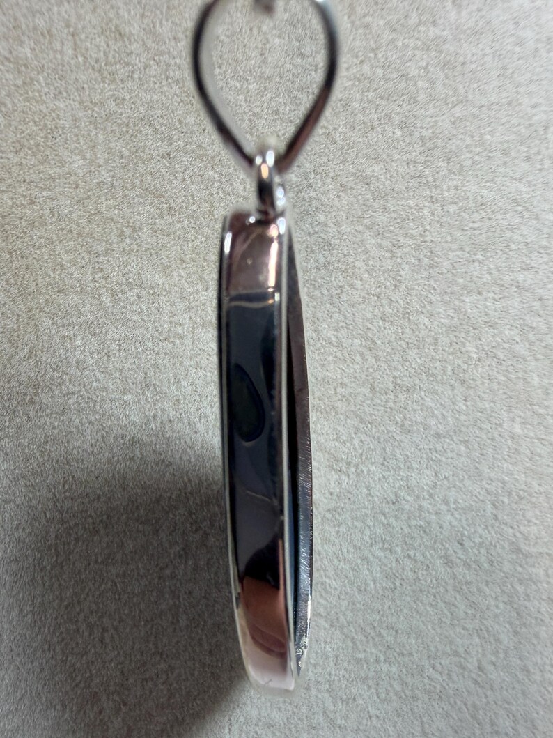 May include: A silver-toned pendant with a dark, reflective center. The pendant has a rounded rectangular shape with a loop at the top for a chain. The edges are polished, creating a contrast with the darker interior.
