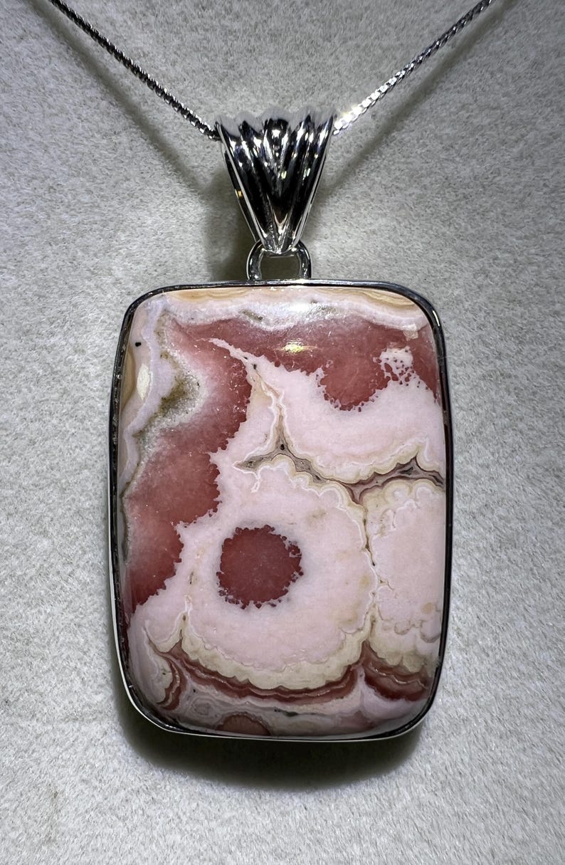 May include: A square pendant necklace featuring a polished stone with a unique pattern of pink, white, and beige swirls. The stone is set in a silver-colored metal frame and hangs from a delicate silver chain.