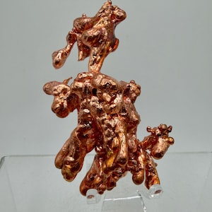 May include: A raw, metallic copper specimen featuring a complex, branching structure with a shiny, reddish-brown surface. The mineral formation displays irregular, bulbous protrusions and a textured, uneven surface resting on a clear stand.