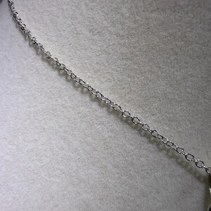 May include: A silver-colored necklace chain with small, evenly spaced links. The chain is displayed against a light gray, textured background, suggesting a jewelry presentation or display.