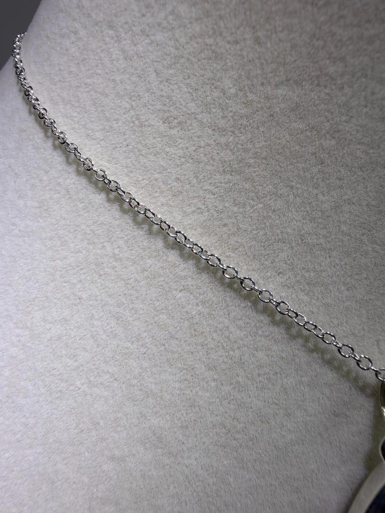 May include: A silver-colored necklace chain with small, evenly spaced links. The chain is displayed against a light gray, textured background, suggesting a jewelry presentation or display.