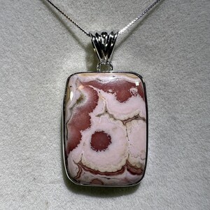 May include: A rectangular pendant necklace featuring a polished stone with a swirling pattern of pink, white, and brown hues. The stone is set in a silver-colored metal frame and hangs from a delicate silver chain.