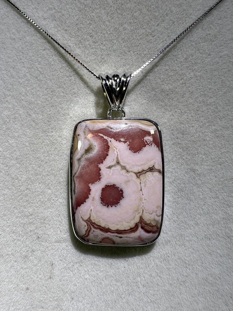 May include: A rectangular pendant necklace featuring a polished stone with a swirling pattern of pink, white, and brown hues. The stone is set in a silver-colored metal frame and hangs from a delicate silver chain.
