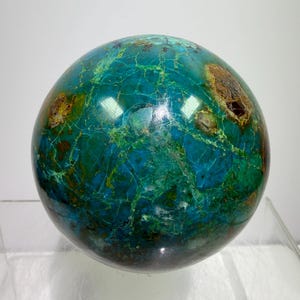 Puede incluir: Una esfera pulida de piedra preciosa de crisocola. La superficie muestra una vibrante mezcla de tonos turquesa, verde azulado y marrón, con intrincados patrones de vetas. La esfera es lisa y reflectante, mostrando la belleza natural de la piedra.