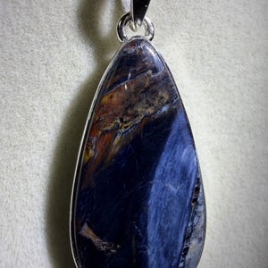 May include: A teardrop-shaped pendant with a polished, dark blue gemstone. The stone has streaks of brown and gold, set in a silver-colored metal frame. The pendant hangs from a silver-colored bail.