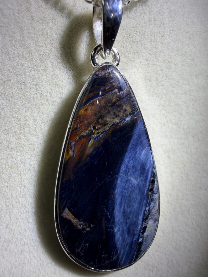 May include: A teardrop-shaped pendant with a polished, dark blue gemstone. The stone has streaks of brown and gold, set in a silver-colored metal frame. The pendant hangs from a silver-colored bail.