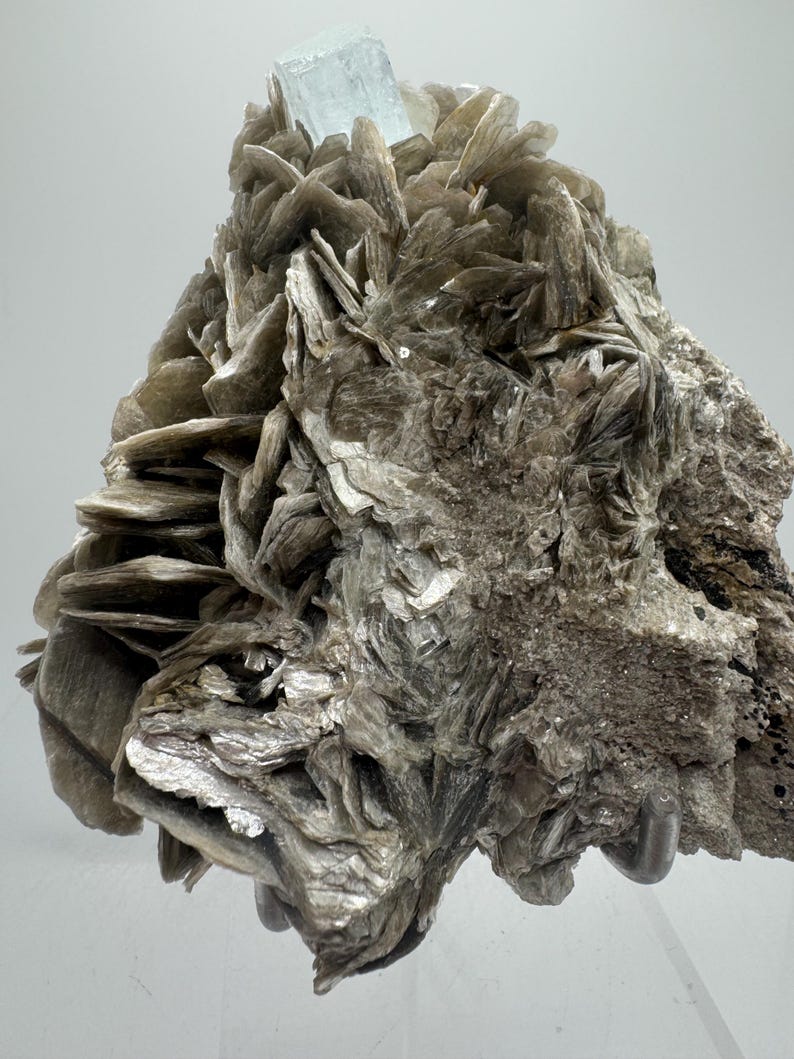May include: A large, gray, rock-like specimen of mica with a clear, light blue aquamarine crystal on top.