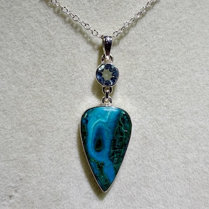 May include: A silver necklace featuring a teardrop-shaped pendant with a vibrant blue and green azurite-malachite stone. Above the main stone, a small, round, faceted light blue gemstone is set in a silver frame, connected to a delicate silver chain.