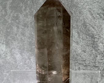 May include: A faceted, smoky quartz crystal point. The crystal is a translucent brown color and has a hexagonal shape with a pointed top. It is displayed on a clear acrylic stand against a textured gray background.