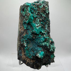 May include: A natural mineral specimen with a dark brown and black base, featuring vibrant, textured turquoise and teal formations. The stone is displayed on a clear acrylic stand, highlighting its unique, organic patterns and textures.