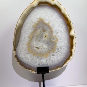 May include: A polished agate slice with a translucent white crystalline center and a tan outer rim, mounted on a black metal stand. The stone features a jagged, natural edge and a textured, sparkling interior with concentric banding patterns.