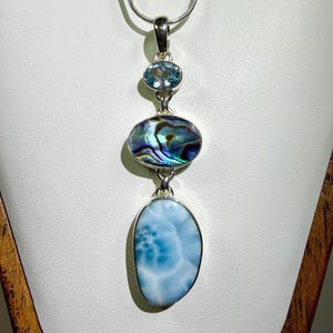 May include: A silver pendant necklace featuring three vertically aligned stones. The top is a small oval light blue gemstone, the middle is an oval iridescent abalone shell, and the bottom is a large, smooth, oval-shaped light blue Larimar stone.
