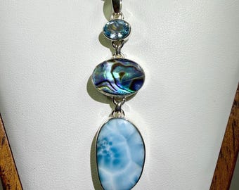 May include: A silver pendant necklace featuring three vertically aligned stones. The top is a small oval light blue gemstone, the middle is an oval iridescent abalone shell, and the bottom is a large, smooth, oval-shaped light blue Larimar stone.