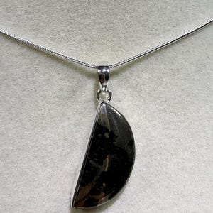 May include: A silver pendant necklace with a half-moon shaped, dark, polished stone. The stone has a black and grey mottled pattern. The pendant hangs from a delicate silver chain, creating a simple and elegant design.