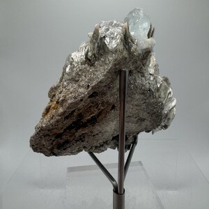 May include: A light blue aquamarine crystal perched atop a bed of silvery mica. The crystal is mounted on a metal stand for display.