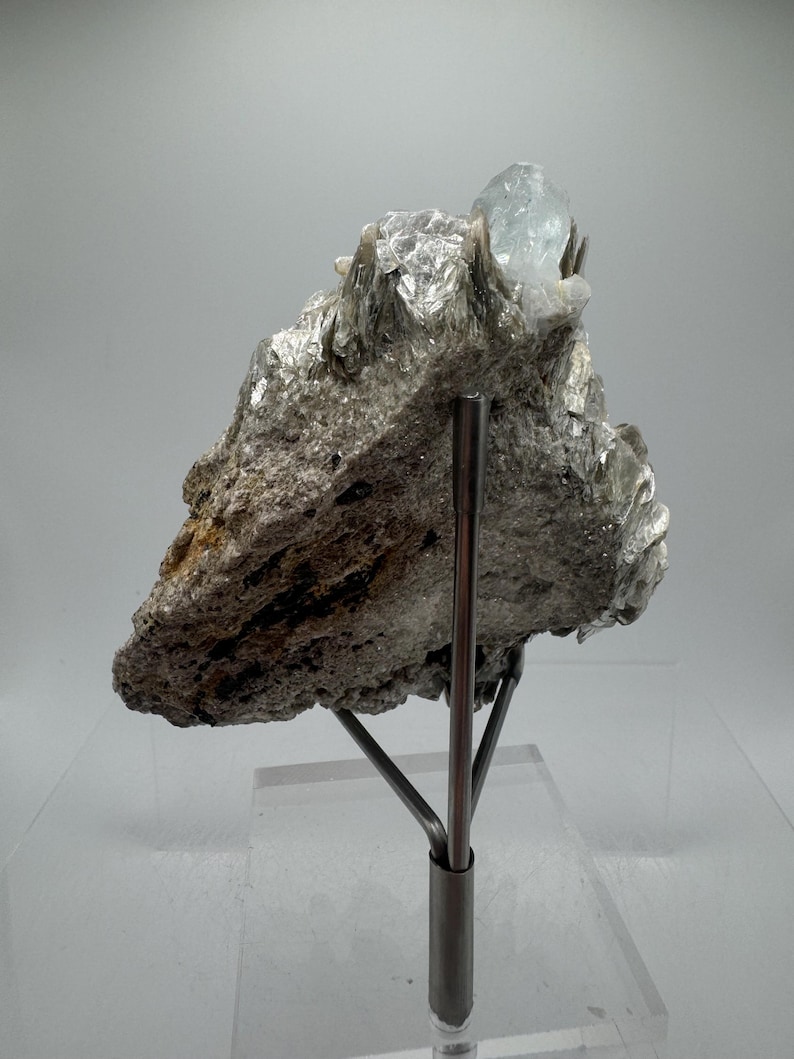 May include: A light blue aquamarine crystal perched atop a bed of silvery mica. The crystal is mounted on a metal stand for display.