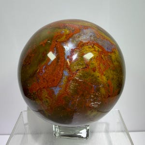 Puede incluir: Una piedra esférica pulida que presenta un patrón complejo de remolinos de color rojo oscuro, amarillo mostaza y morado apagado. La superficie es lisa y reflectante, mostrando inclusiones minerales naturales y transiciones de color.