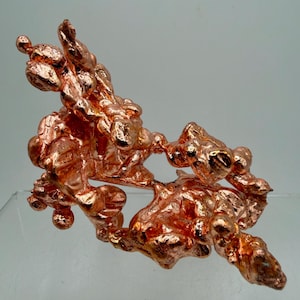 May include: A raw, metallic copper specimen featuring a complex, branching structure with numerous rounded, bulbous protrusions. The surface displays a bright, reflective reddish-orange luster with varied textures, creating an organic, irregular form.
