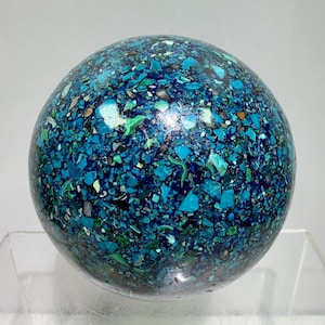 Puede incluir: Un objeto esférico que presenta un mosaico de fragmentos de piedra triturada en varios tonos de azul, turquesa y verde oscuro. La superficie está pulida con un acabado liso y brillante, mostrando un patrón denso y multicolor de piezas angulares.