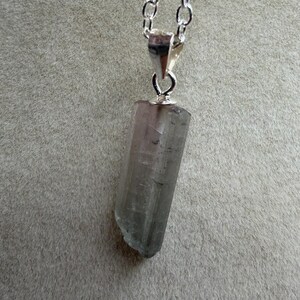 May include: A silver chain necklace with a pendant featuring a raw, light grey crystal. The crystal is a long, rectangular shape with a slightly rough, uneven edge. The chain is made of small, linked silver pieces.
