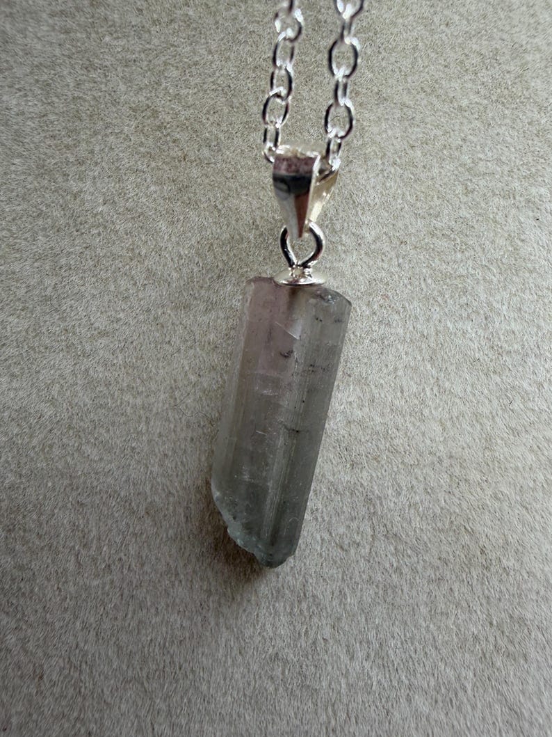 May include: A silver chain necklace with a pendant featuring a raw, light grey crystal. The crystal is a long, rectangular shape with a slightly rough, uneven edge. The chain is made of small, linked silver pieces.