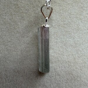 May include: A pendant necklace featuring a long, rectangular, semi-translucent crystal with a colour gradient transitioning from a pale pinkish-purple at the top to a muted greyish-green at the base, suspended from a silver-toned metal chain.