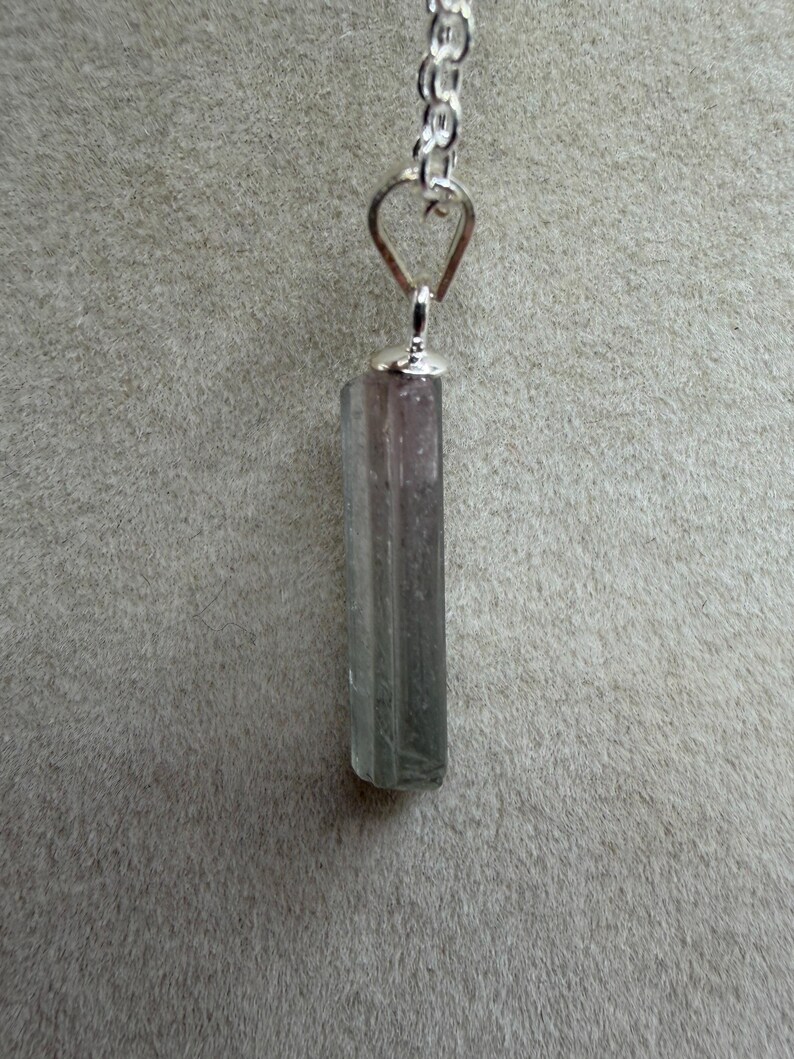 May include: A pendant necklace featuring a long, rectangular, semi-translucent crystal with a colour gradient transitioning from a pale pinkish-purple at the top to a muted greyish-green at the base, suspended from a silver-toned metal chain.