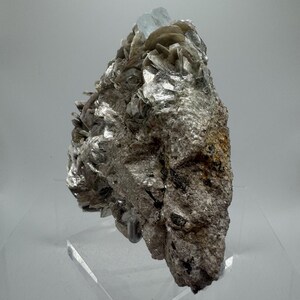 May include: A cluster of light blue aquamarine crystals embedded in a matrix of gray and brown rock. The rock is on a clear acrylic display stand.