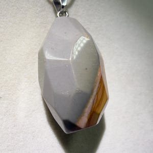 May include: A faceted, gray gemstone pendant with a silver-colored bail hangs from a silver chain. The stone has a mix of gray, brown, and pink hues. The pendant is a geometric shape with multiple facets.