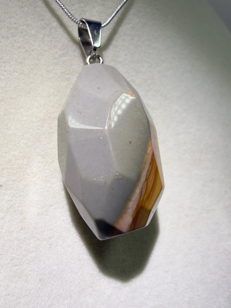May include: A faceted, gray gemstone pendant with a silver-colored bail hangs from a silver chain. The stone has a mix of gray, brown, and pink hues. The pendant is a geometric shape with multiple facets.