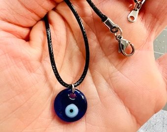 Pode incluir: Um colar de cordão preto com um pingente de olho turco azul. O pingente é redondo e tem um olho branco com uma pupila preta.