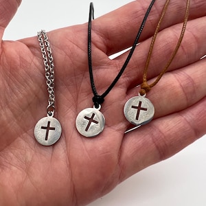 May include: Three cross pendant necklaces. One has a silver chain, the others have black and brown cords. Each pendant is a silver-colored disc with a cross cutout. The necklaces are displayed on a light background.