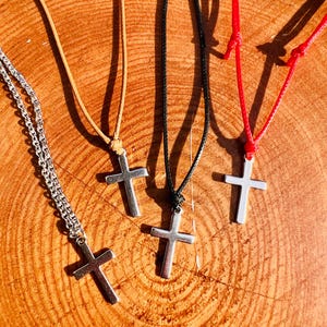 May include: A collection of silver cross pendants on various cords. The necklaces include a silver chain, a tan leather cord, a black cord, and a red cord. The crosses are all the same size and shape, with a simple, classic design.