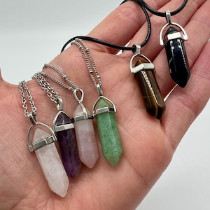 May include: A selection of gemstone pendant necklaces in shades of clear, purple, pink, green, brown, and black. Each pendant is a polished, elongated crystal with a silver-tone metal cap and chain or cord. The necklaces are presented against a plain backdrop.