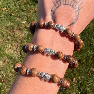 May include: Three wooden bead bracelets with silver elephant charms. The beads are a warm brown color with a wood grain pattern. The bracelets are worn on a wrist against a background of green grass.