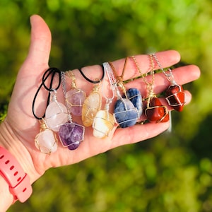 Puede incluir: Una colección de colgantes de piedras preciosas, cada uno con una piedra única de forma irregular envuelta en alambre. Las piedras varían en color, incluyendo rosa, morado, amarillo, azul y rojo. Los collares tienen diferentes estilos de cadena.