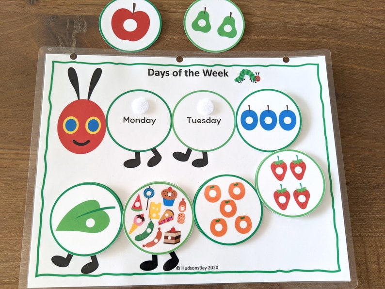 May include: A colorful educational toy featuring a caterpillar with different days of the week on each segment. The toy includes various images, such as an apple, a pear, a leaf, and a plate of food. The text "Days of the Week" is printed at the top of the toy.