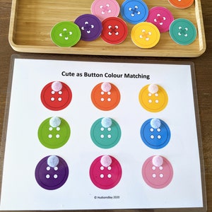 May include: A printable activity sheet with the text "Cute as Button Colour Matching". The sheet has nine circles with different colored buttons inside. Each circle has a white pom-pom on top.