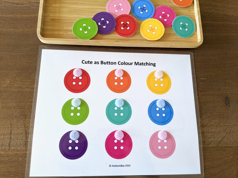 May include: A printable activity sheet with the text "Cute as Button Colour Matching". The sheet has nine circles with different colored buttons inside. Each circle has a white pom-pom on top.