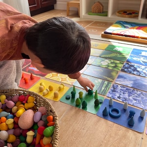 May include: A child plays with colorful wooden blocks and matching cards. The cards feature images of nature, such as trees, flowers, and blueberries. The blocks are in a basket with other wooden toys.