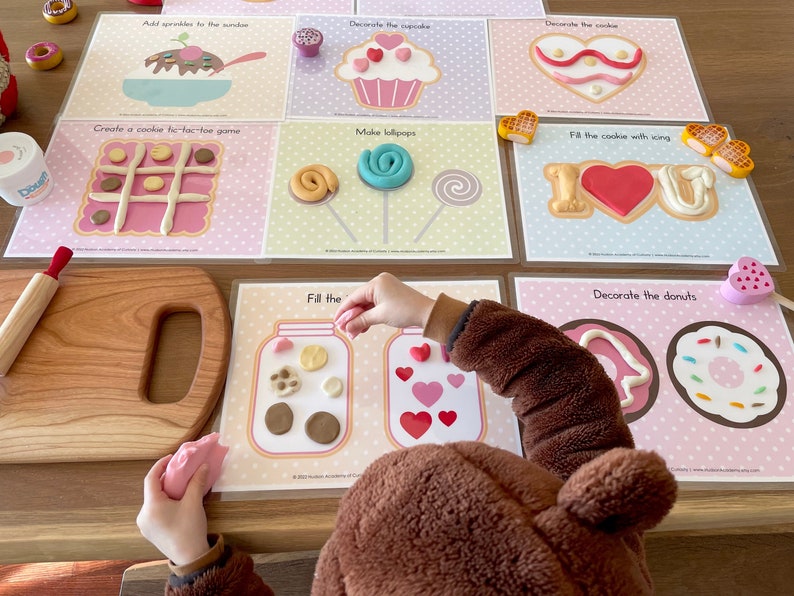 May include: A set of eight printable play dough activity cards with a pink and white polka dot background. The cards feature various activities such as decorating a cupcake, cookie, and donuts, making lollipops, and creating a tic-tac-toe game. The cards have the text 'Decorate the cupcake', 'Decorate the cookie', 'Create a cookie tic-tac-toe game', 'Make lollipops', 'Fill the cookie with icing', 'Fill the', 'Decorate the donuts'.