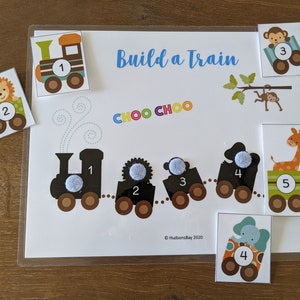 May include: A colorful train building activity with a black and white train engine, four train cars, and five animal-themed cards. The cards feature a lion, monkey, giraffe, elephant, and a train engine. The cards have numbers 1 through 5 on them. The text "Build a Train" and "CHOO CHOO" is on the activity sheet. &copy; Hudson's Bay 2020 is in the bottom right corner.