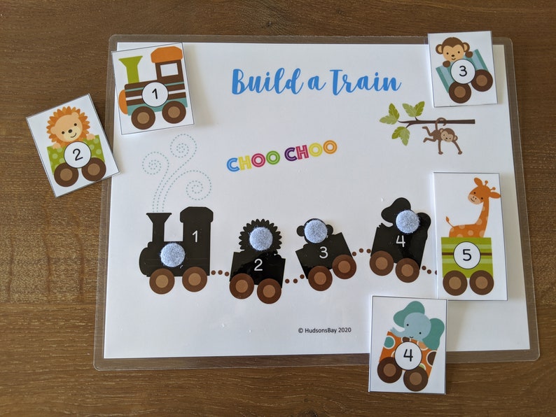 May include: A colorful train building activity with a black and white train engine, four train cars, and five animal-themed cards. The cards feature a lion, monkey, giraffe, elephant, and a train engine. The cards have numbers 1 through 5 on them. The text "Build a Train" and "CHOO CHOO" is on the activity sheet. &copy; Hudson's Bay 2020 is in the bottom right corner.