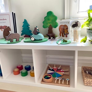 May include: A white shelf unit with three compartments. The top shelf has a brown bear busy book, wooden bear figurines, a wooden car, a wooden tree, and a color wheel game. The middle shelf has a rainbow wooden puzzle and a set of rainbow stacking rings. The bottom shelf has a color wheel game and a wicker basket with a stuffed animal inside.