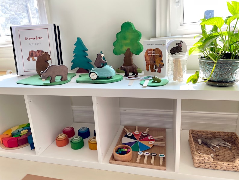 May include: A white shelf unit with three compartments. The top shelf has a brown bear busy book, wooden bear figurines, a wooden car, a wooden tree, and a color wheel game. The middle shelf has a rainbow wooden puzzle and a set of rainbow stacking rings. The bottom shelf has a color wheel game and a wicker basket with a stuffed animal inside.