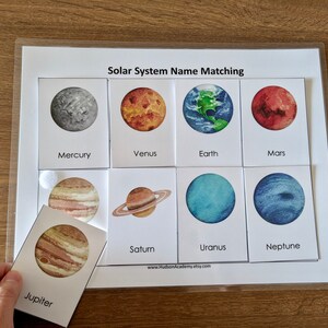 May include: A solar system name matching game with colorful illustrations of planets. The game includes cards with images of planets and corresponding names, such as Mercury, Venus, Earth, Mars, Jupiter, Saturn, Uranus, and Neptune.