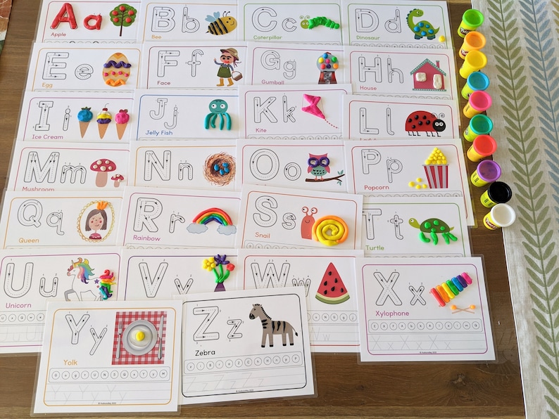 May include: A set of 26 alphabet cards with colorful playdough shapes representing each letter. The cards feature dotted lines for tracing and a word for each letter. The cards are laid out on a wooden table with containers of playdough.